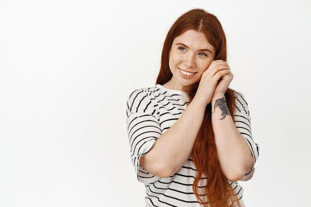 El retrato de una pelirroja soñadora tiene pensamientos románticos, mirando con admiración y cara feliz, sonriendo, tomándose de la mano cerca de la piel natural limpia con pecas, fondo blanco