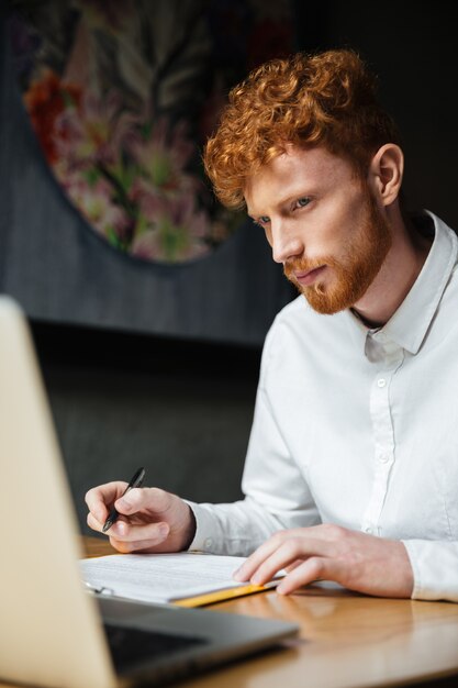 Retrato de pensar hombre pelirrojo joven mirando portátil en el lugar de trabajo