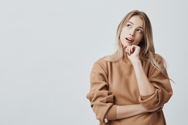 Retrato de pensativa mujer rubia de ensueño y de ensueño imagina algo en su mente, mira hacia arriba, sumida en pensamientos, tocando la barbilla. Atractiva joven rubia sueña con los fines de semana