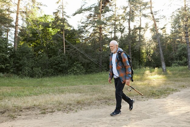 Retrato de pensionista masculino europeo calvo sin afeitar con mochila, llevando caña de pescar o carrete giratorio, yendo a pescar en la orilla del río. Pesca recreativa, estilo de vida activo y saludable y ocio.