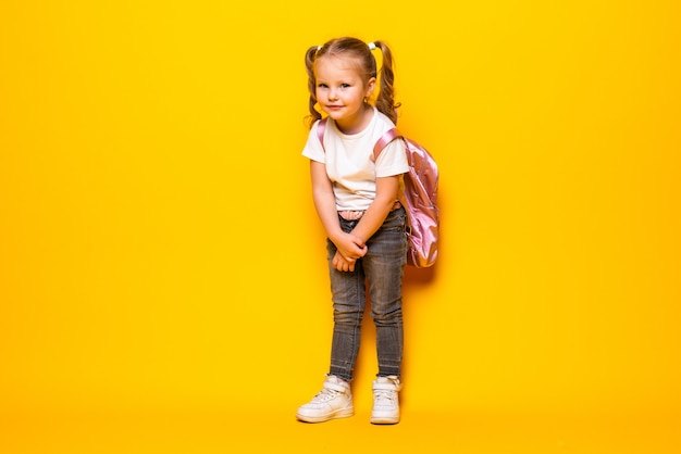 Retrato de una pequeña colegiala sonriente con mochila en la pared amarilla