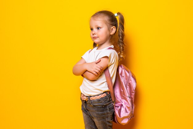 Retrato de una pequeña colegiala sonriente con mochila en la pared amarilla