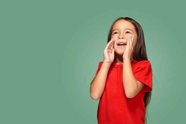 Retrato de una pequeña niña bonita de pie de lado y llamando a alguien sosteniendo su mano cerca de su boca con una camiseta roja, sobre un fondo verde