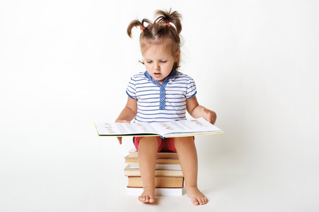 Foto gratuita el retrato del pequeño niño adorable se sienta en la pila de libros, sostiene un libro interesante, ve imágenes, trata de leer algunas palabras, se prepara para la escuela, aislado en blanco. niña inteligente