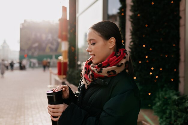 El retrato de perfil de una chica morena con una cálida chaqueta oscura y una bufanda disfruta de un café y camina al aire libre a la luz del sol y en un cálido día de invierno