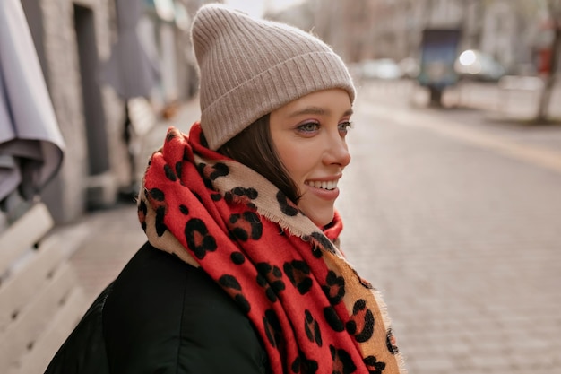 Foto gratuita el retrato de perfil de una elegante dama sonriente con gorro de punto y bufanda colorida está sentado al aire libre en un banco y esperando amigos en la ciudad