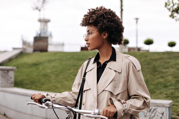 Retrato de perfil de hermosa mujer morena de piel oscura posando con bicicleta Encantadora dama rizada con anteojos y gabardina beige camina afuera