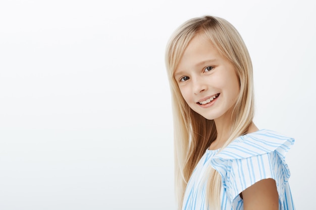 Retrato de perfil interior de una encantadora joven caucásica de pelo rubio con una moderna blusa azul, sonriendo ampliamente y sintiéndose feliz después de la lección de piano, relajándose y jugando con sus compañeros de clase sobre una pared gris