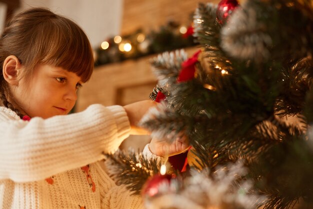 Retrato de perfil de niña caucásica decorando el árbol de Navidad, vestido de suéter blanco, con cabello oscuro, esperando la víspera de año nuevo, estando de humor festivo, Feliz Navidad.