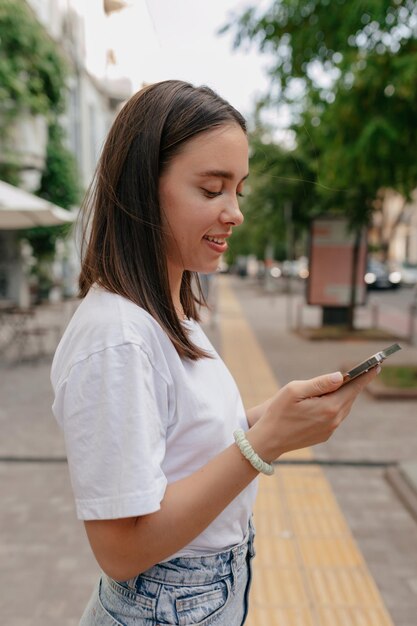 Retrato de perfil de niña feliz sonriente en camiseta blanca está desplazando el teléfono inteligente en la ciudad en un cálido día de primavera Una niña encantadora está caminando en la ciudad