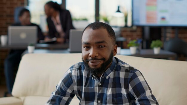 Retrato de una persona afroamericana sonriendo en la oficina, sentada en el sofá para trabajar en una laptop. Empleado de la empresa que utiliza estadísticas financieras para crear desarrollo laboral y crecimiento empresarial.