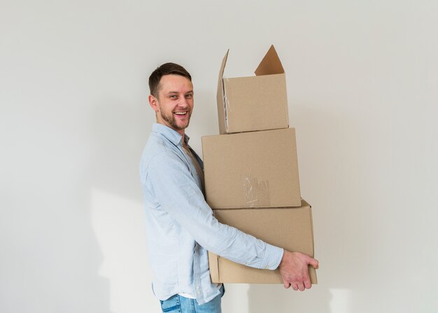 Retrato de una pila que lleva sonriente del hombre joven de cajas de cartón contra la pared blanca