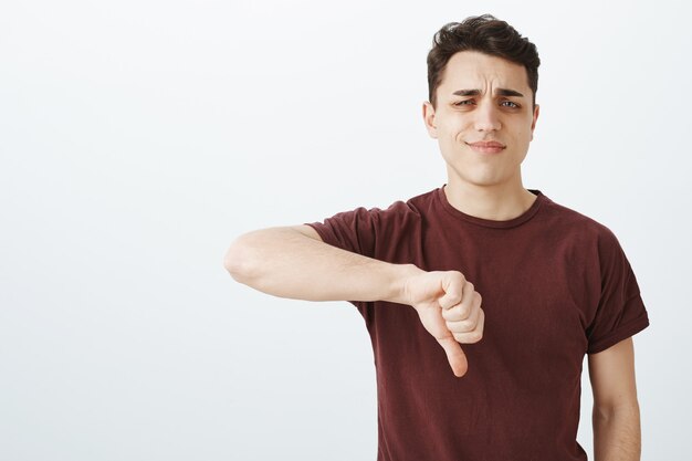 Retrato de poco impresionado disgustado atractivo estudiante masculino en camiseta con los pulgares hacia abajo