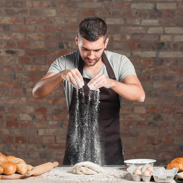 El retrato del polvo del panadero de sexo masculino amasa la masa con la harina en el contador de cocina