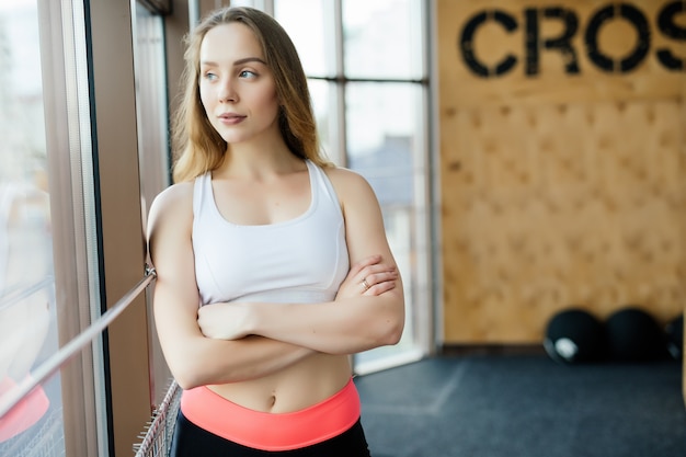 Retrato de positividad y niña sonriendo a la cámara y posando con los brazos cruzados en el gimnasio. Chica seductora con ropa deportiva blanca, top y leggins. fondo de madera