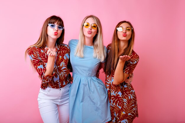 Retrato positivo brillante de tres feliz hipster mujer divirtiéndose y enviando besos al aire, ropa de verano a juego de colores y gafas de sol coloridas,