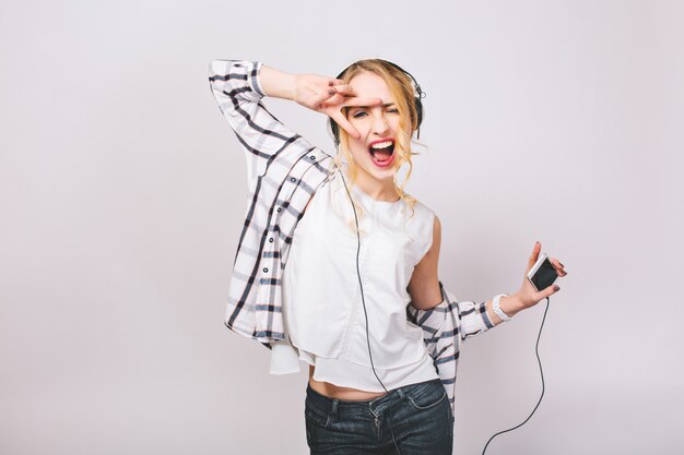 Retrato positivo de la energía alegre chica con cabello rubio en traje casual escuchando música con auriculares grandes. Ella está bailando y sosteniendo un teléfono inteligente. Aislado.