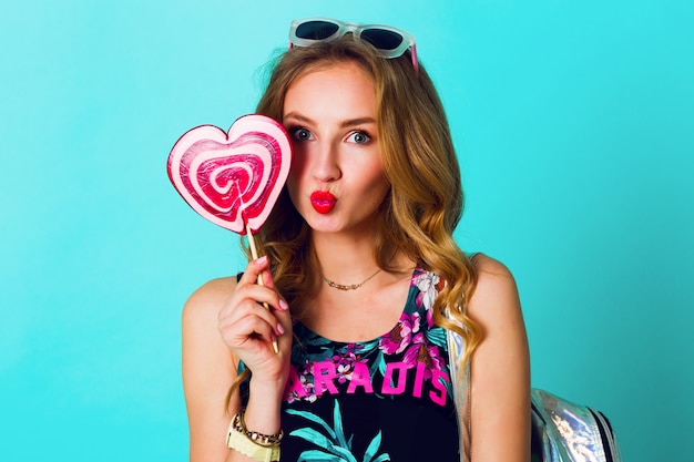 Retrato positivo del estudio de la mujer loca de la moda divertida joven atractiva que presenta en fondo de la pared azul en traje del estilo del verano con la piruleta rosada que lleva la tapa de la impresión, la mochila de neón y las gafas lindas.