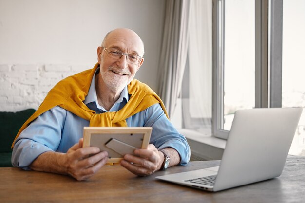 Retrato de positivo exitoso empleado senior atractivo con barba gris trabajando en el interior de la oficina moderna, usando la computadora portátil, sosteniendo el marco de fotos y sonriendo mientras extraña a sus nietos
