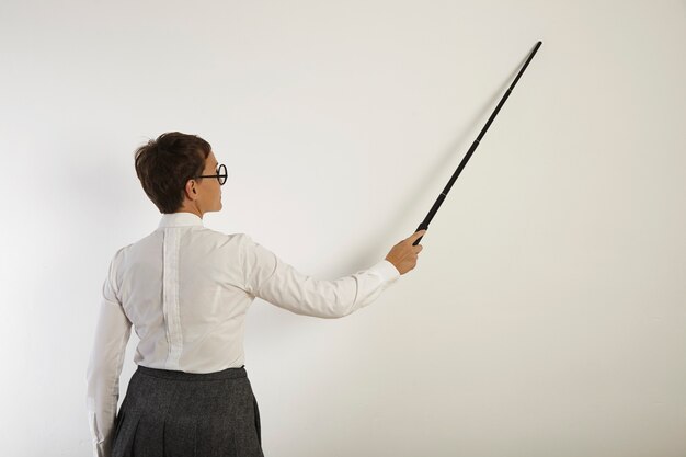 Retrato posterior de una profesora blanca de aspecto serio en blusa, falda y gafas apuntando a una pared blanca con un puntero negro