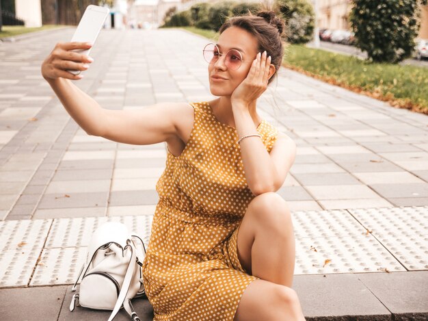 Retrato del primer de la muchacha morena sonriente hermosa en vestido amarillo del inconformista del verano. Modelo tomando selfie en teléfono inteligente. Mujer haciendo fotos en un día cálido y soleado en la calle con gafas de sol