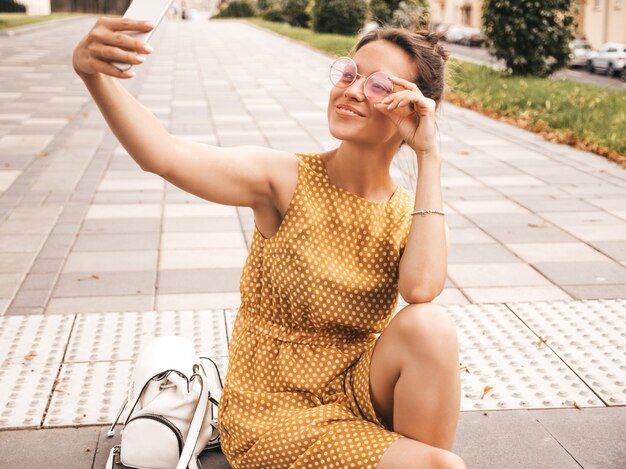 Retrato del primer de la muchacha morena sonriente hermosa en vestido amarillo del inconformista del verano. Modelo tomando selfie en teléfono inteligente. Mujer haciendo fotos en un día cálido y soleado en la calle con gafas de sol