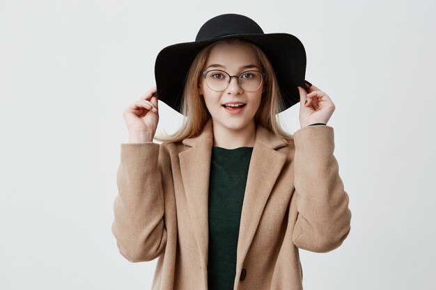 Retrato del primer de la muchacha rubia joven con la piel pura, las lentes y la sonrisa que llevan el sombrero negro y la capa aislados. Linda mujer disfrutando de su estilo.