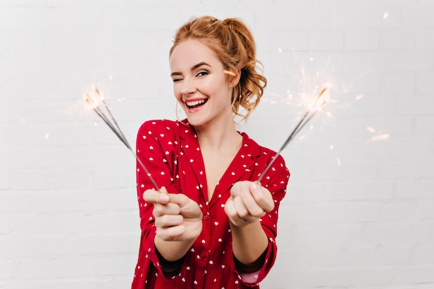 Retrato de primer plano de la adorable niña sonriente celebrando el año nuevo con luces de bengala. mujer elegante en pijama rojo divertido esperando la Navidad.