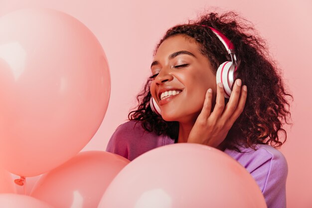 Retrato de primer plano de una agradable niña africana en auriculares disfrutando de la fiesta. Alegre mujer negra escuchando música mientras celebraba un cumpleaños.