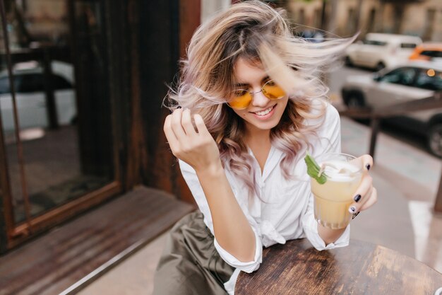 Retrato de primer plano al aire libre de dama adorable con una sonrisa suave disfrutando de un café helado en la cafetería