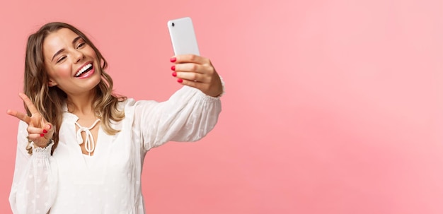 Retrato de primer plano de una alegre y encantadora chica rubia femenina con vestido blanco que se toma selfie en el teléfono móvil hace un signo de paz kawaii mientras toma una foto capturando el fondo rosa del momento primaveral