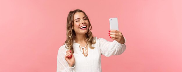 Retrato de primer plano de una alegre y optimista chica rubia sonriente con un vestido blanco riéndose como un amigo grabando videollamadas en una aplicación móvil tomando una foto selfie en el fondo rosa del teléfono inteligente