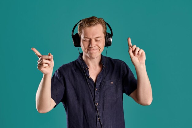 Retrato de primer plano de un apuesto hombre adulto de jengibre con una elegante camiseta azul marino escuchando la música a través de unos auriculares mientras posa en el fondo azul del estudio. Expresiones faciales humanas. emociones sinceras