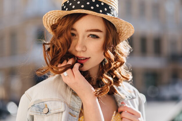 Retrato de primer plano de atractiva mujer blanca con sombrero posando en la calle. Tiro al aire libre de chica jengibre interesada en chaqueta vaquera.