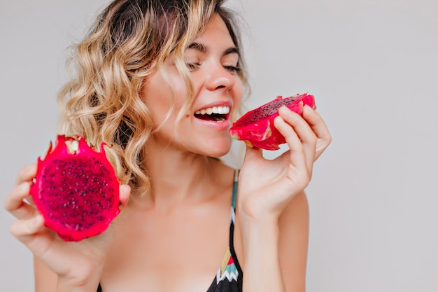 Retrato de primer plano de atractiva mujer bronceada con peinado corto comiendo fruta del dragón. Chica refinada disfrutando de una jugosa pitaya roja.