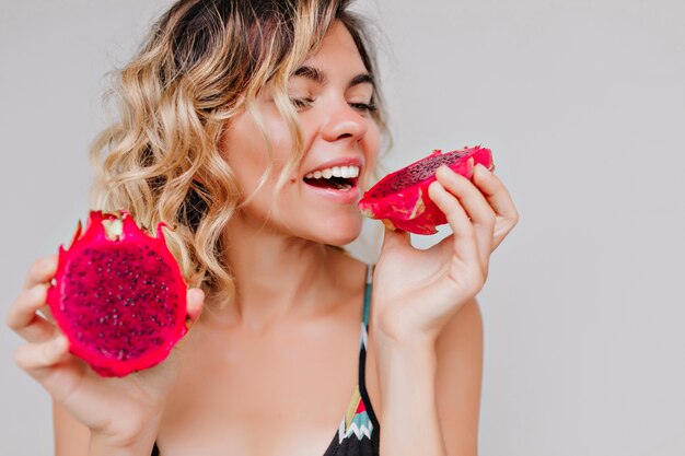 Retrato de primer plano de atractiva mujer bronceada con peinado corto comiendo fruta del dragón. Chica refinada disfrutando de una jugosa pitaya roja.