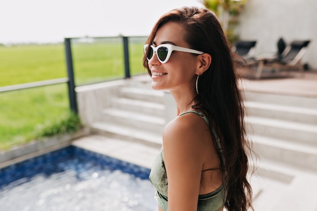 Retrato de primer plano de atractiva mujer sonriente feliz en gafas y traje de baño posando durante la sesión de fotos por campo verde y piscina. chica relajante en balneario en buen día soleado