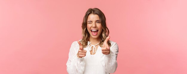 Retrato de primer plano de una atrevida y despreocupada guapa emotiva chica rubia señalando pistolas con el dedo a la cámara mientras pide unirse a su equipo para convertirse en miembro invitando a probar el producto usted mismo sonriendo descarado