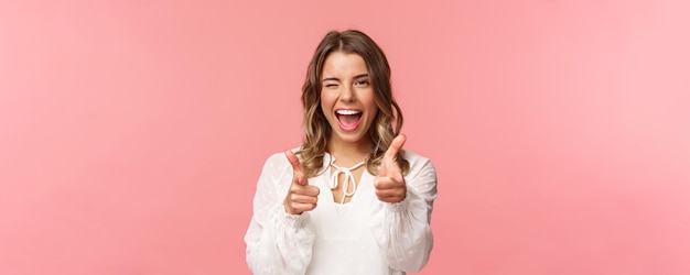 Retrato de primer plano de una atrevida y despreocupada guapa emotiva chica rubia señalando pistolas con el dedo a la cámara mientras pide unirse a su equipo para convertirse en miembro invitando a probar el producto usted mismo sonriendo descarado