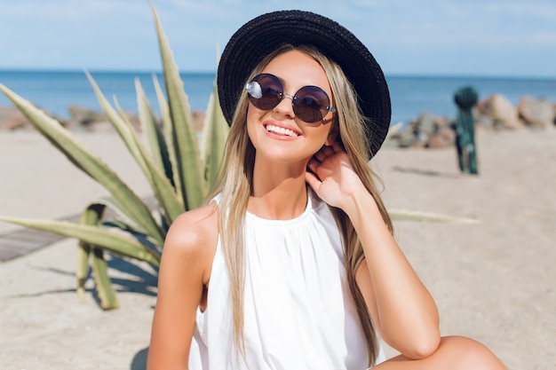 Retrato de primer plano de una chica bastante rubia con el pelo largo está sentado en la playa cerca del mar. Ella sonríe a la cámara.