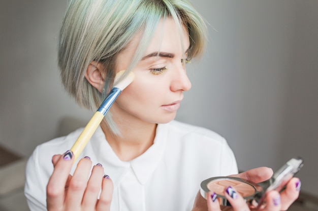 Retrato de primer plano de una chica bonita con pelo corto blanco sobre fondo gris. Lleva un vestido blanco, se mira al espejo en la mano y se empolva la cara.