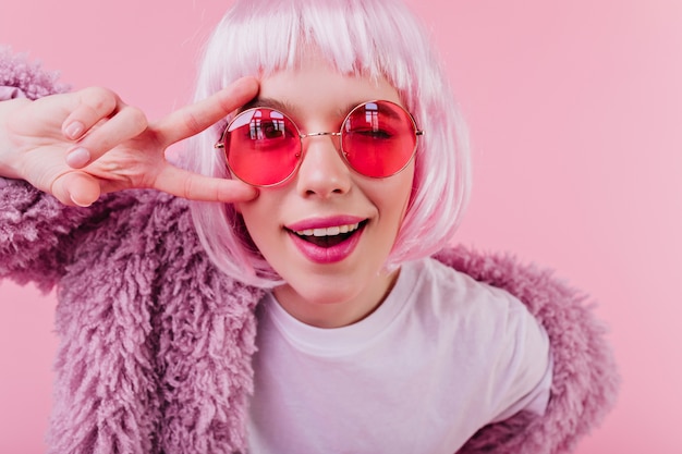 Retrato de primer plano de chica elegante en moda peruke. filmación en interiores de risa joven con gafas de sol aislado en la pared rosa