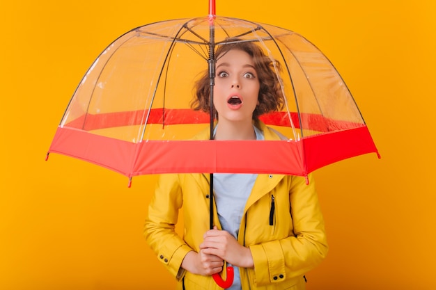 Retrato de primer plano de una chica entusiasta con peinado rizado de pie bajo la sombrilla. Fotografía interior de modelo femenino molesto en impermeable con paraguas.