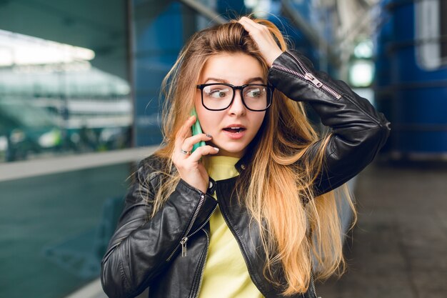 Retrato de primer plano de una chica guapa de pie afuera en el aeropuerto. Ella tiene cabello largo y chaqueta negra. Ella está hablando por teléfono. Ella se ve maravillada.