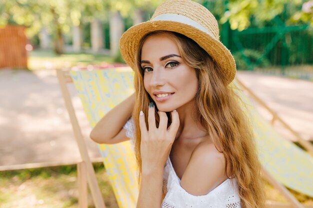 Retrato de primer plano de chica inspirada con piel ligeramente bronceada jugando con su largo cabello dorado. Foto al aire libre de una mujer joven sonriente en un canotier vintage y un vestido blanco de verano.