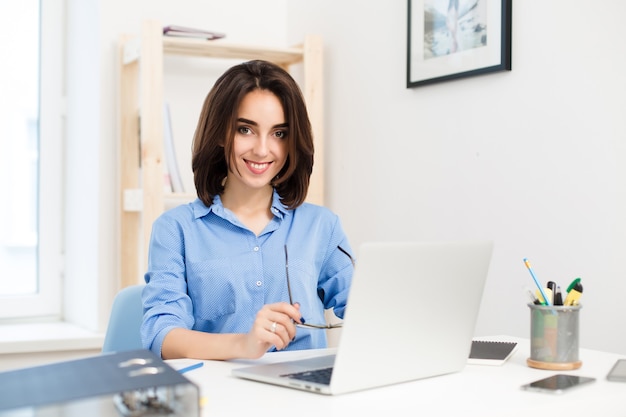 Retrato de primer plano de una chica joven y bonita con una camisa azul. Ella está sentada a la mesa en la oficina y sonriendo a la cámara.