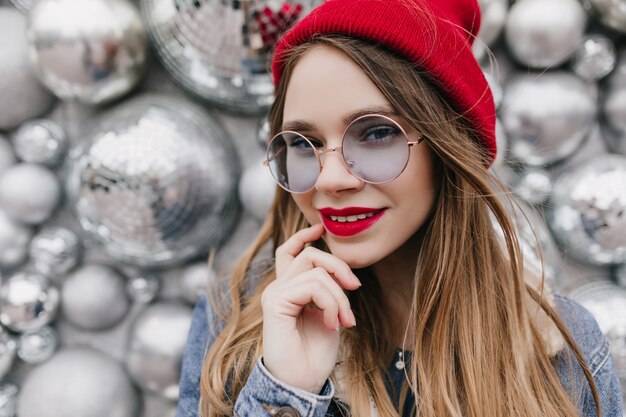 Retrato de primer plano de una chica maravillosa con maquillaje brillante posando en la pared de brillo. Foto de modelo de mujer extática lleva sombrero rojo y gafas azules redondas.