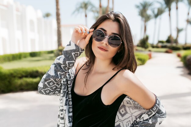 Retrato de primer plano de la chica de moda con gafas de sol y camisa elegante, posando en el parque y disfrutando del sol del sur en vacaciones. Increíble mujer joven con cabello lacio oscuro de pie afuera.