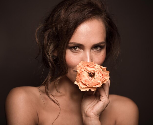 Retrato de primer plano de una chica modelo profesional sonriendo aislada en la oscuridad Hermosa dama con cabello castaño mirando a la cámara y sosteniendo una rosa naranja cerca de su boca