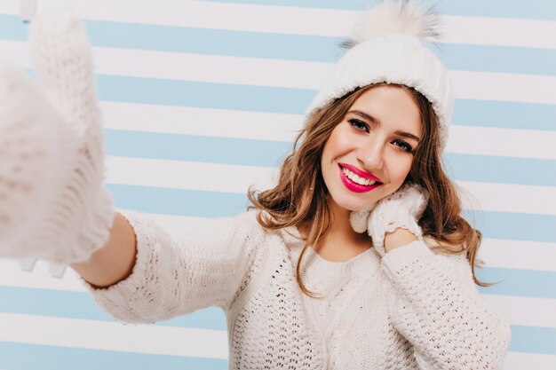 Retrato de primer plano de chica morena positiva en lindos guantes de lana haciendo selfie en pared rayada. Señora riendo con sombrero blanco y guantes de punto tomando una foto de sí misma.